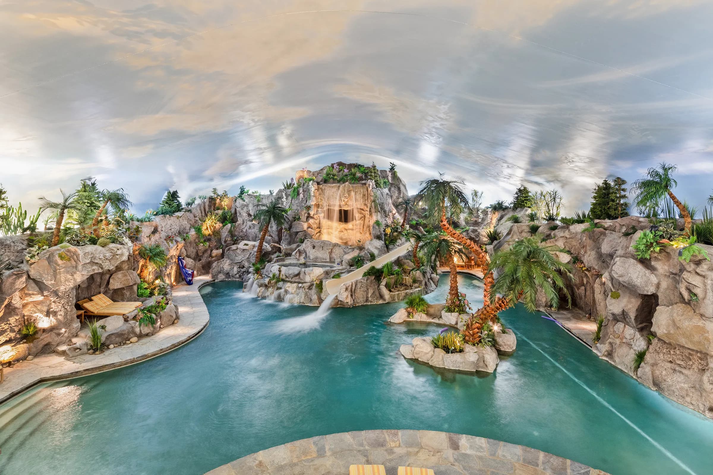 The indoor water park at Juggler Meadow: a palm-lined lagoon with stone grottos, a waterfall, and a water slide beneath a painted sky