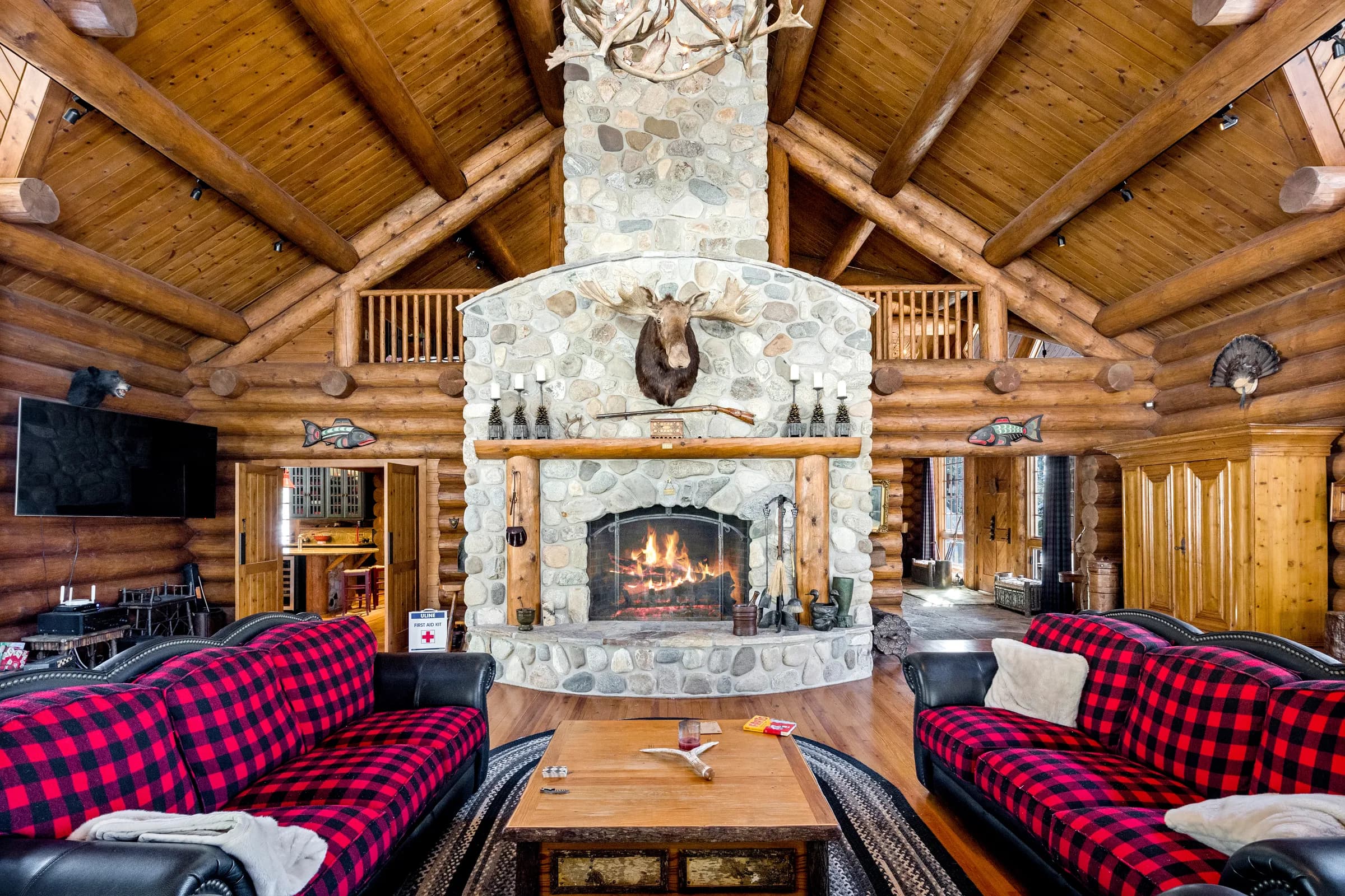 The great room's two-story river-stone fireplace, fire lit beneath the moose mount, flanked by buffalo-plaid sofas
